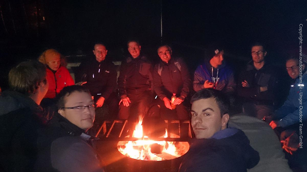 Firmenevent Survivalkurs - nach einem Tag voller Aktivitäten sitzt die Gruppe am späten Abend entspannt am wärmenden Lagerfeuer.